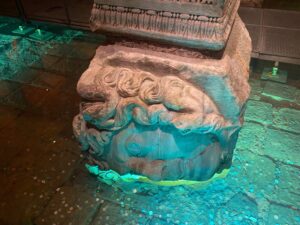 Sideways Medusa head carved on a column base in the Basilica Cistern in Istanbul, lit in teal light with coins on the floor around it
