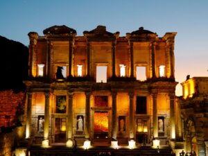 Library of Celsus in Ephesus lit by golden floodlights against a deep blue evening sky, with small silhouettes of visitors at its base