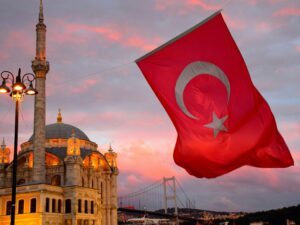 Large Turkish flag with a white crescent and star waving in a pink sunset sky beside the Ortaköy Mosque and the Bosphorus Bridge in Istanbul