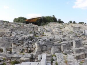 Stone walls and ruined houses of the ancient city of Troy at Hisarlık, with a protective roof over the dig in the distance