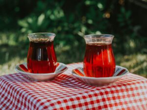 Two glasses of Turkish tea on saucers on a red and white checkered tablecloth outdoors, with green leaves behind